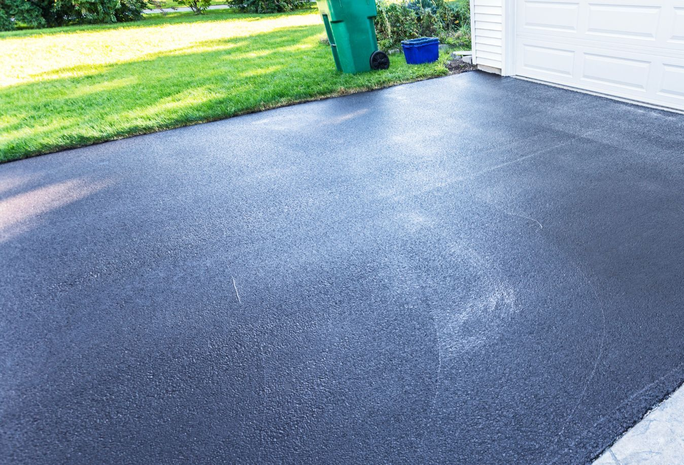 Freshly sealed black driveway beside a white garage and green lawn