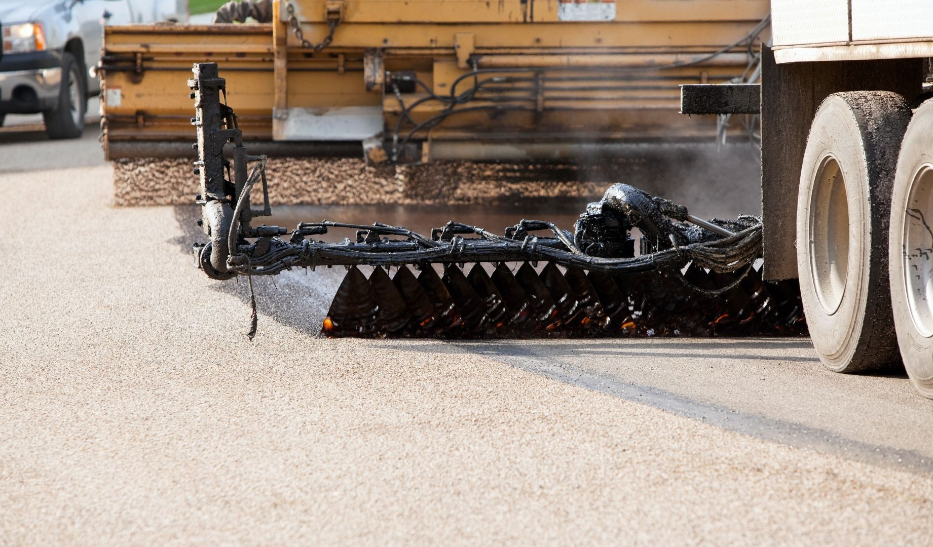 Road construction machine spraying fresh asphalt on a street beside a truck wheel