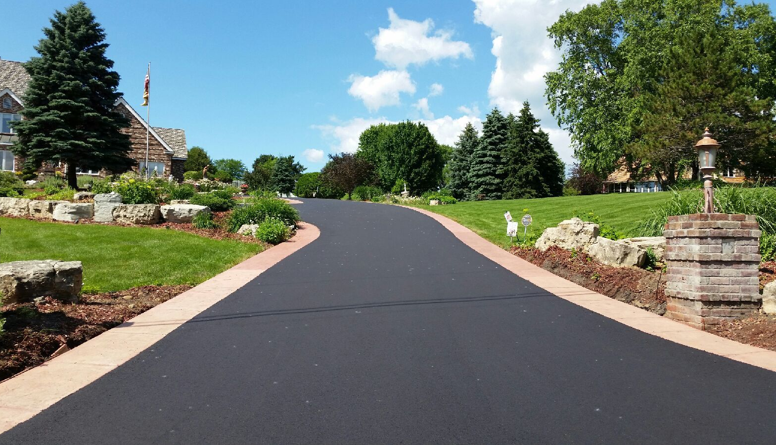 Curved paved driveway lined with flowers and greenery leading to houses under a blue sky