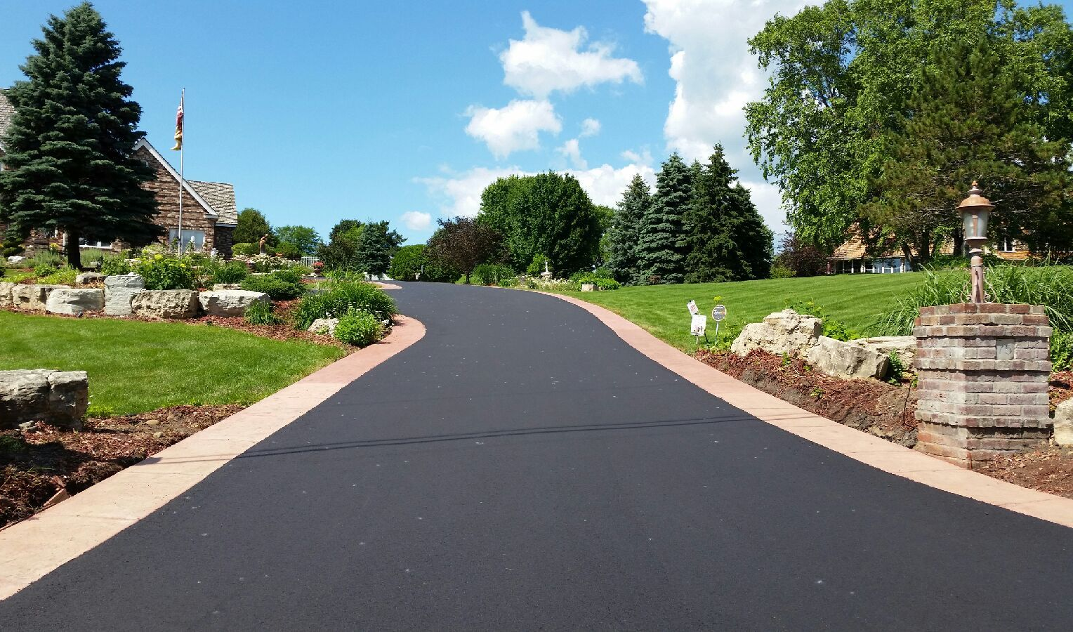 Curved asphalt driveway through landscaped garden with trees, flowers, and blue sky