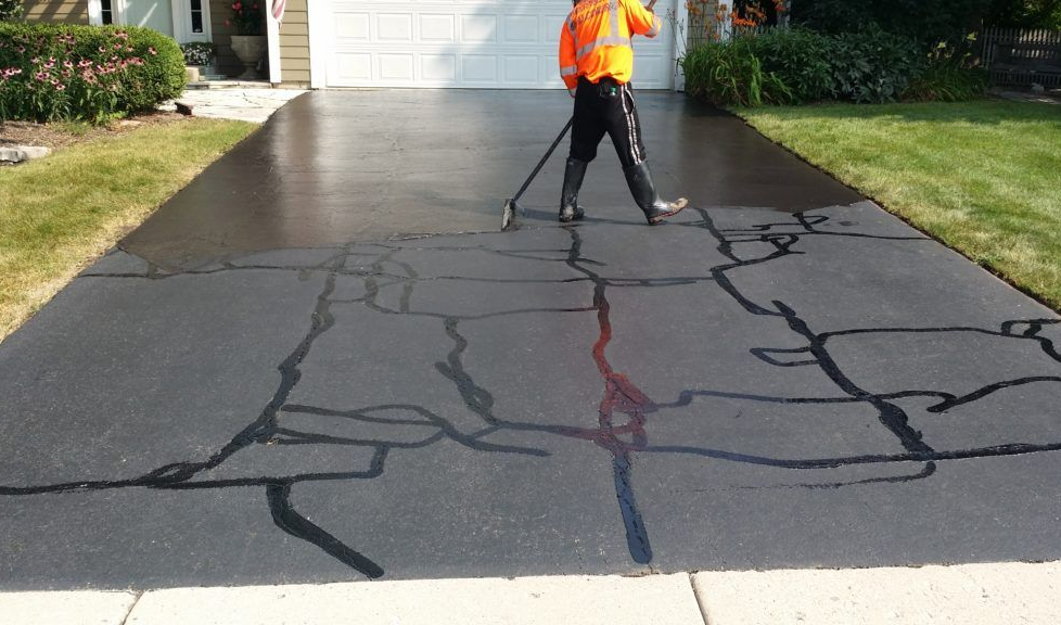 Worker spray-sealing a cracked asphalt driveway with a black coating.