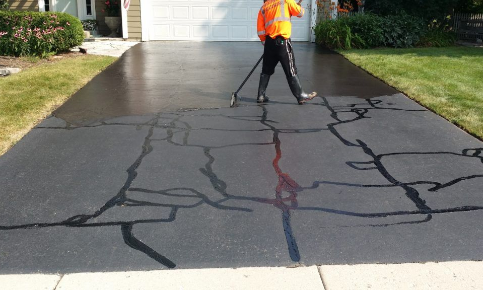 Worker sealing cracks in a black asphalt driveway with a long-handled tool