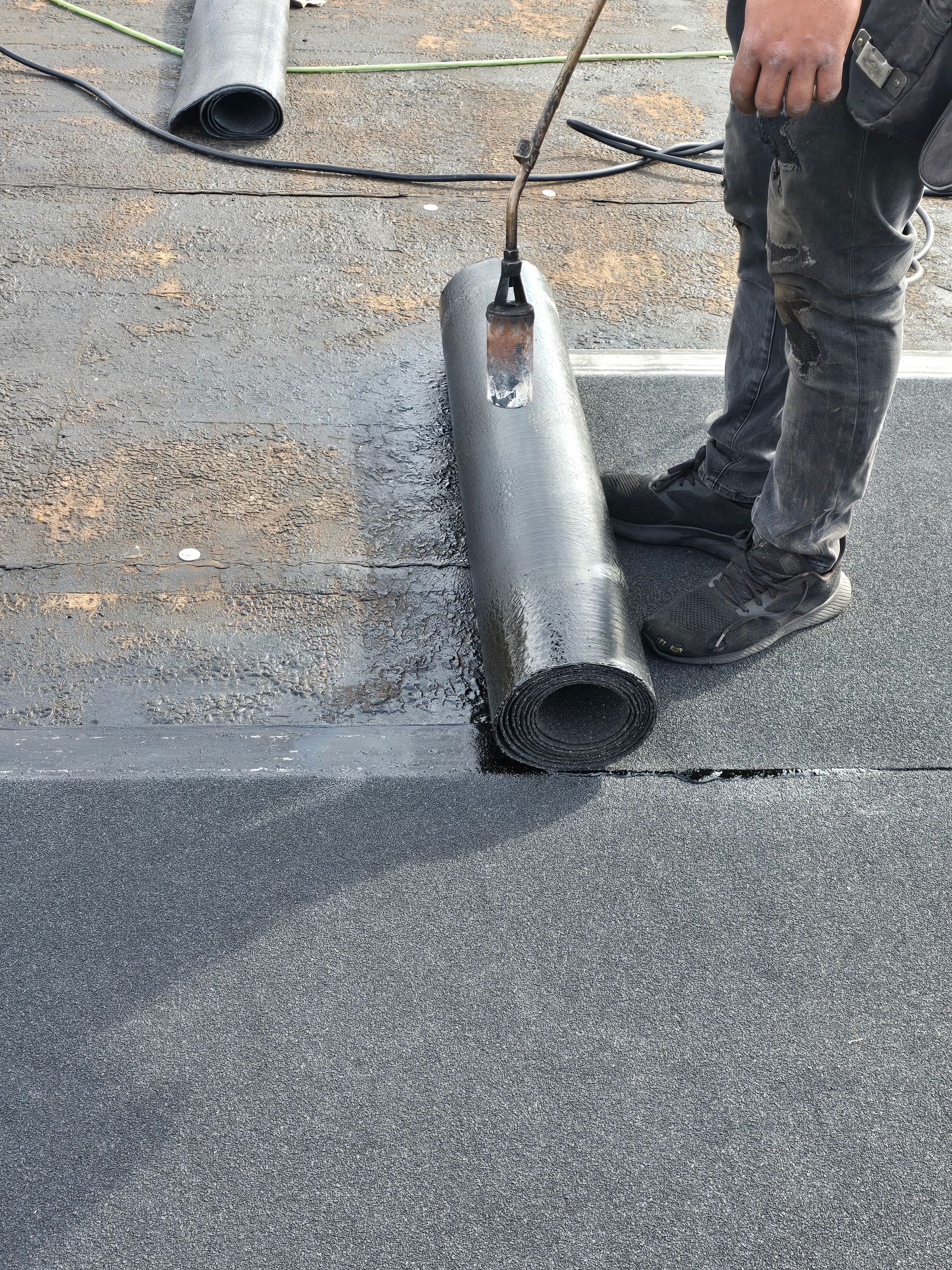 Worker using a torch to heat rolled roofing on a flat roof, with a second roll nearby