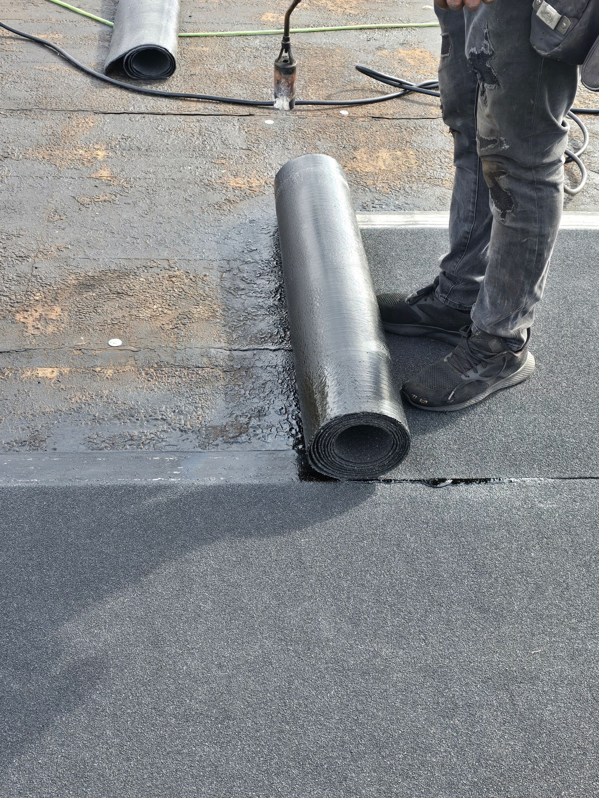 Rolled roofing membrane on gravel roof beside a worker’s boots