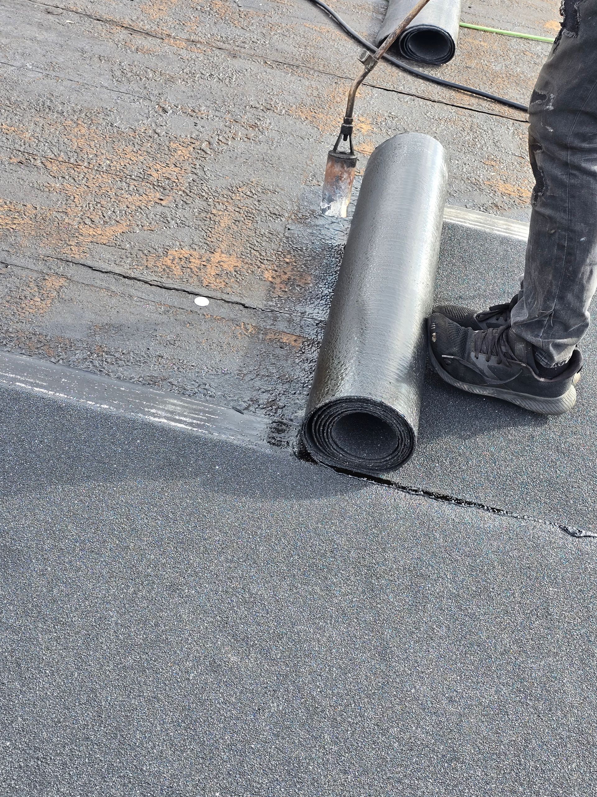 Worker installing a rolled roofing membrane on a flat roof with a torch tool