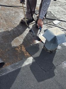 Person repairing a roof vent on a gravel rooftop, with tools and black roofing material nearby