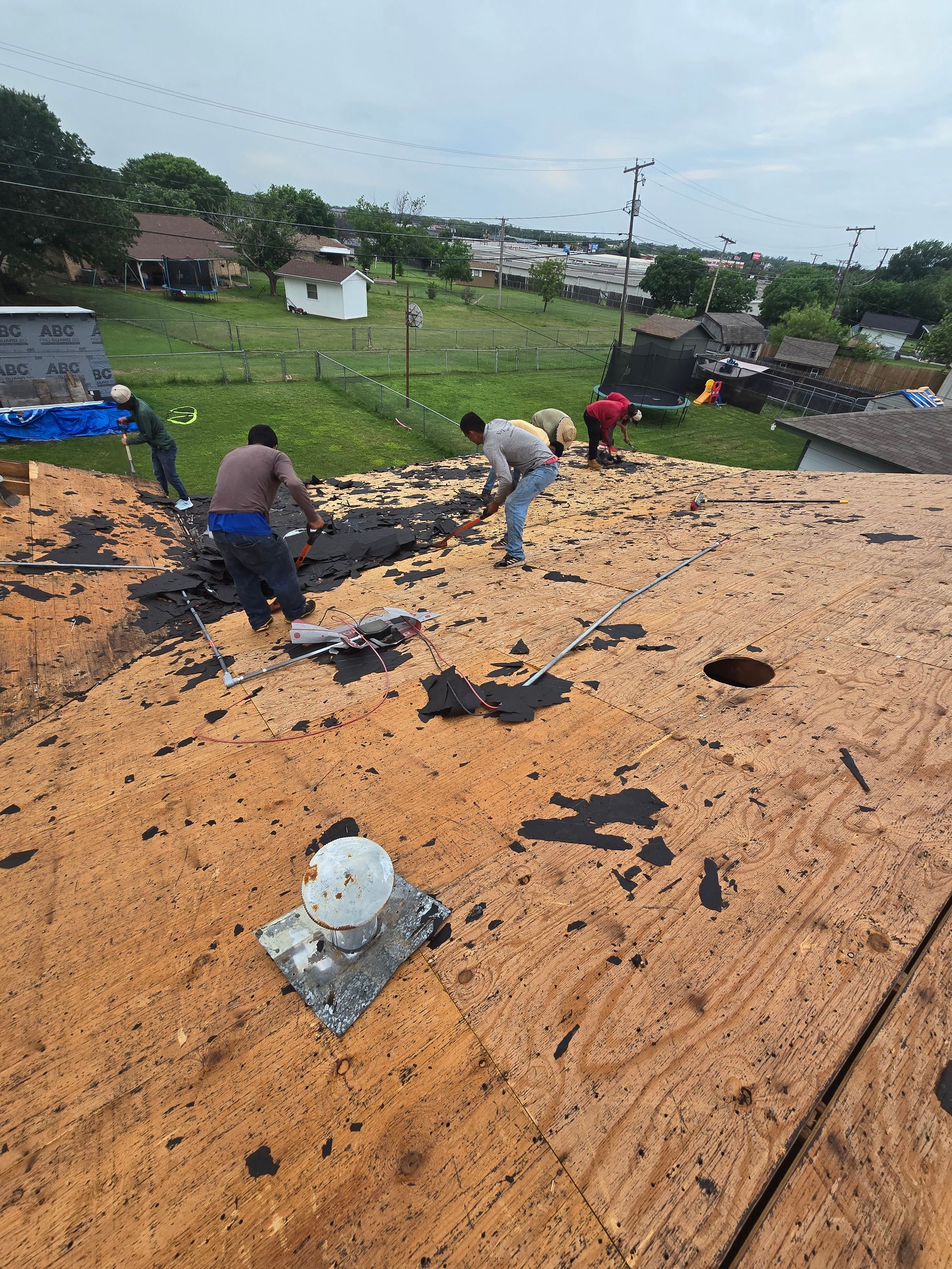 Workers on a roof clearing debris after storm damage, with a grassy neighborhood in the background.