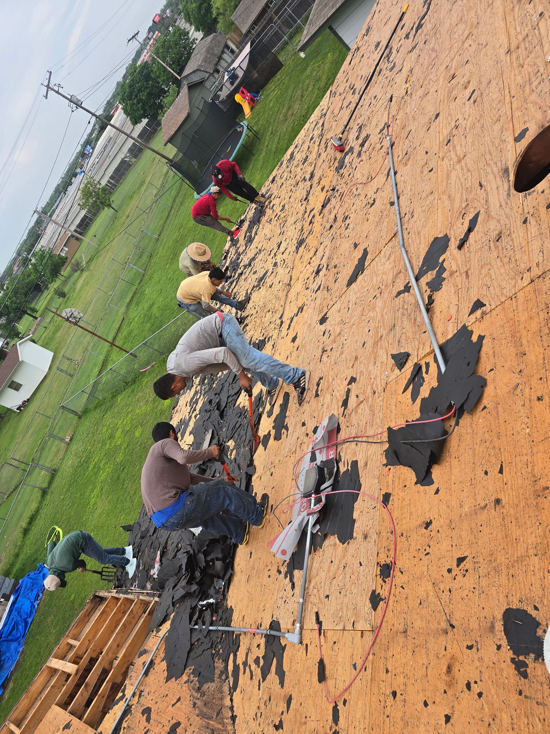 Workers on a roof installing shingles, with stacks of roofing materials on a rainy day.