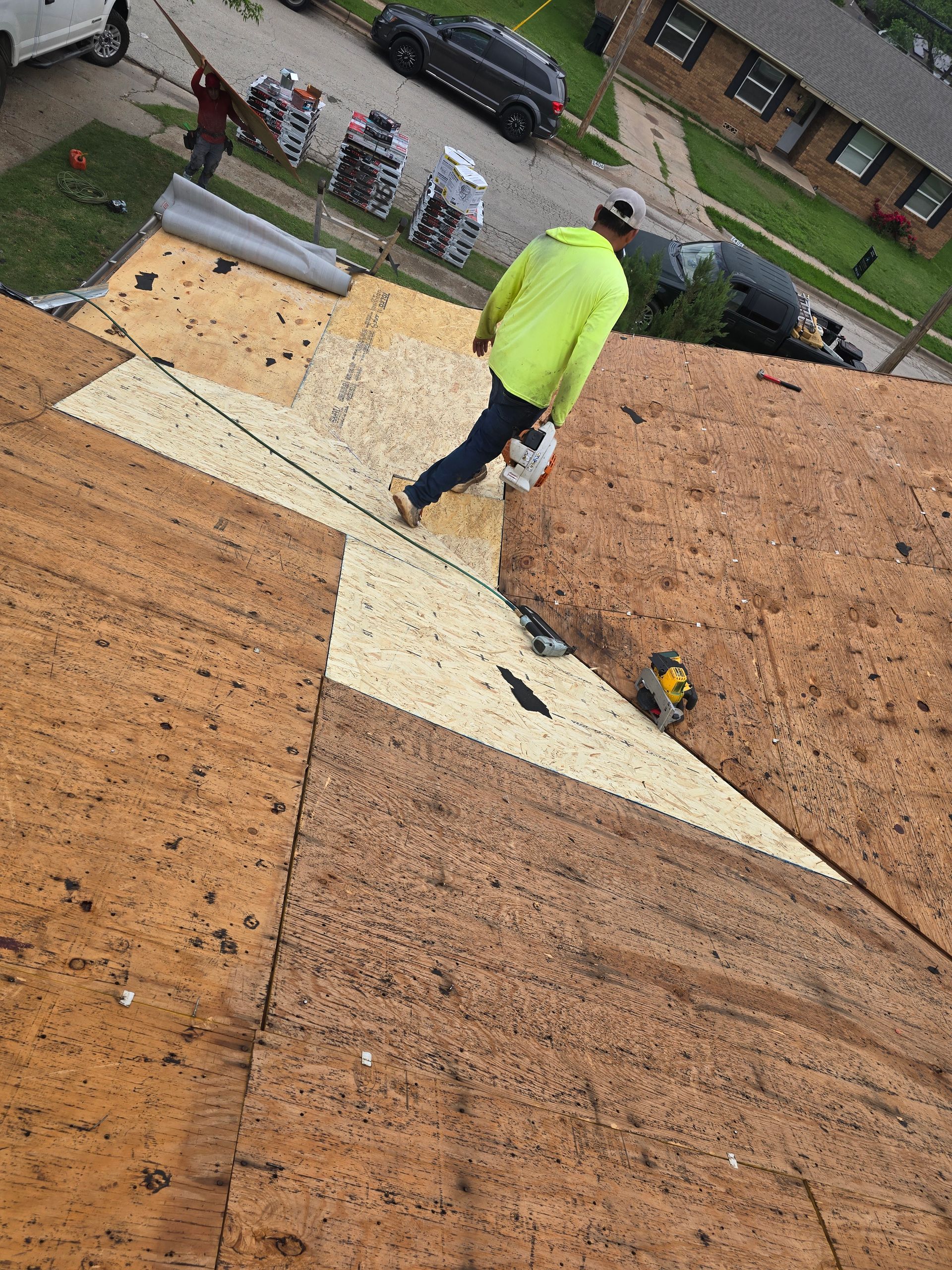 Worker on a roof installing a light plywood patch between brown shingles, with tools nearby and cars below.