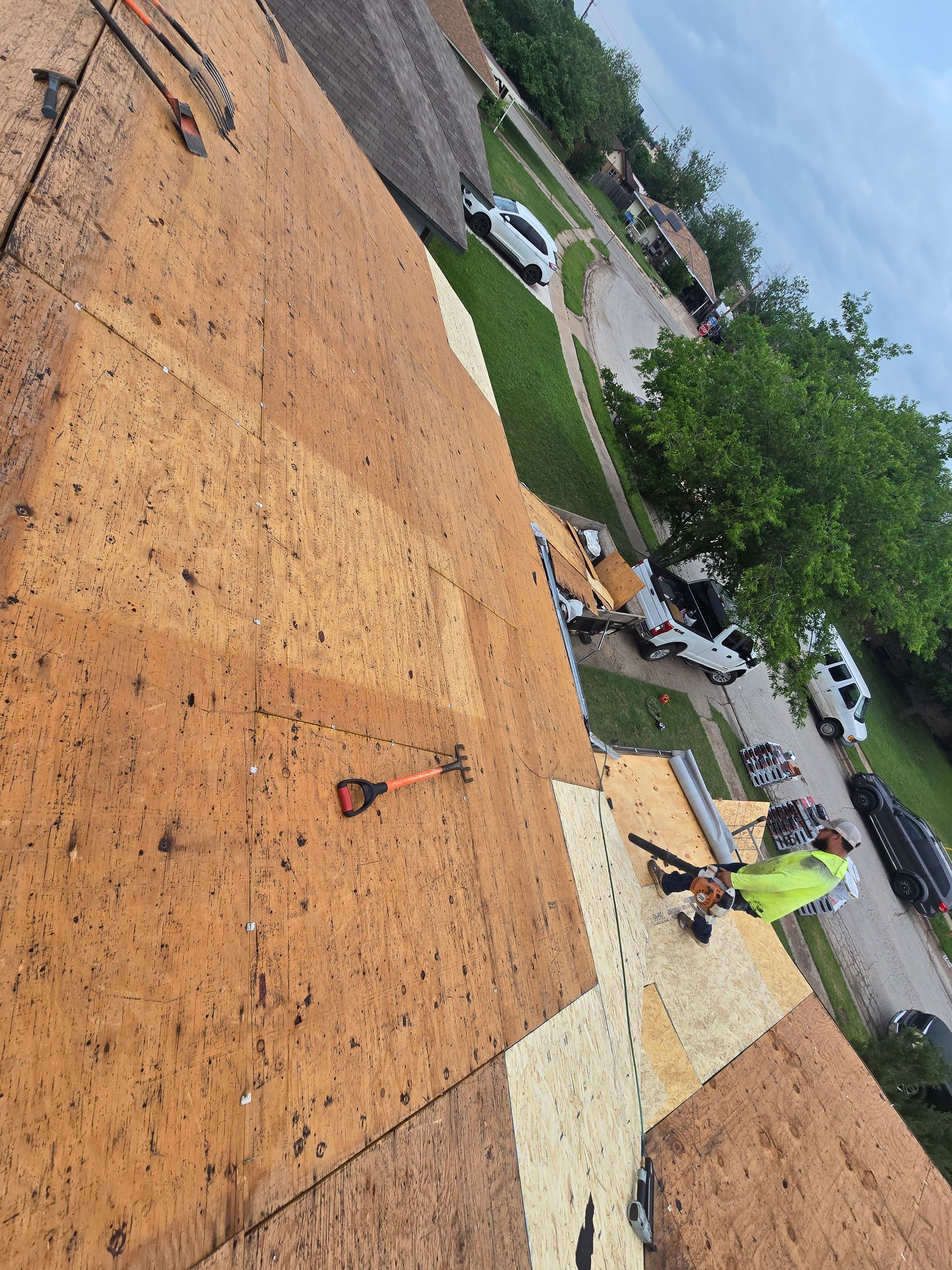 Roof workers on an orange shingle roof, one in a neon vest near white underlayment, with trees and parked cars nearby