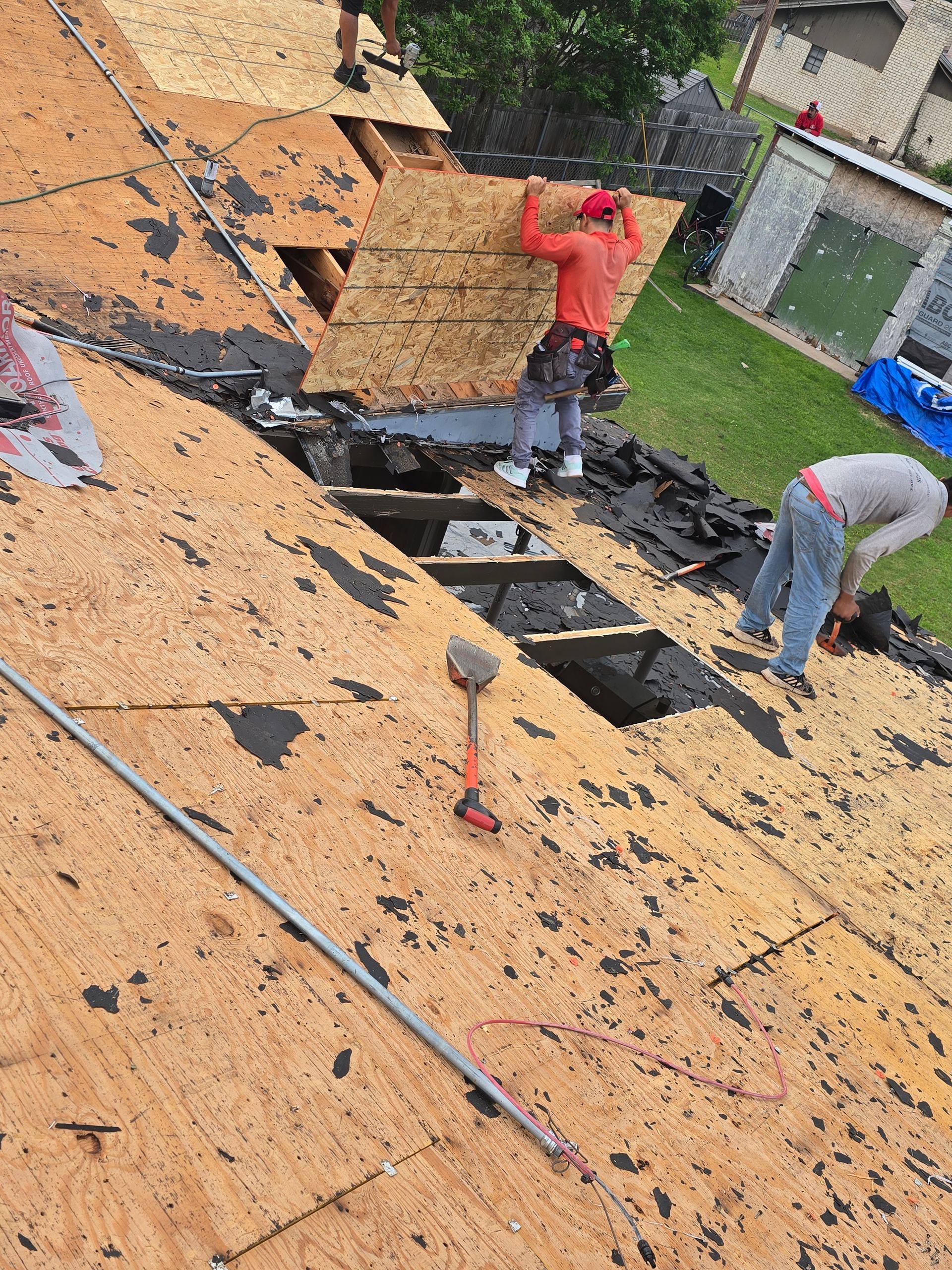 Roofers removing shingles on a damaged roof, with one in red and another in gray working near a dark exposed section