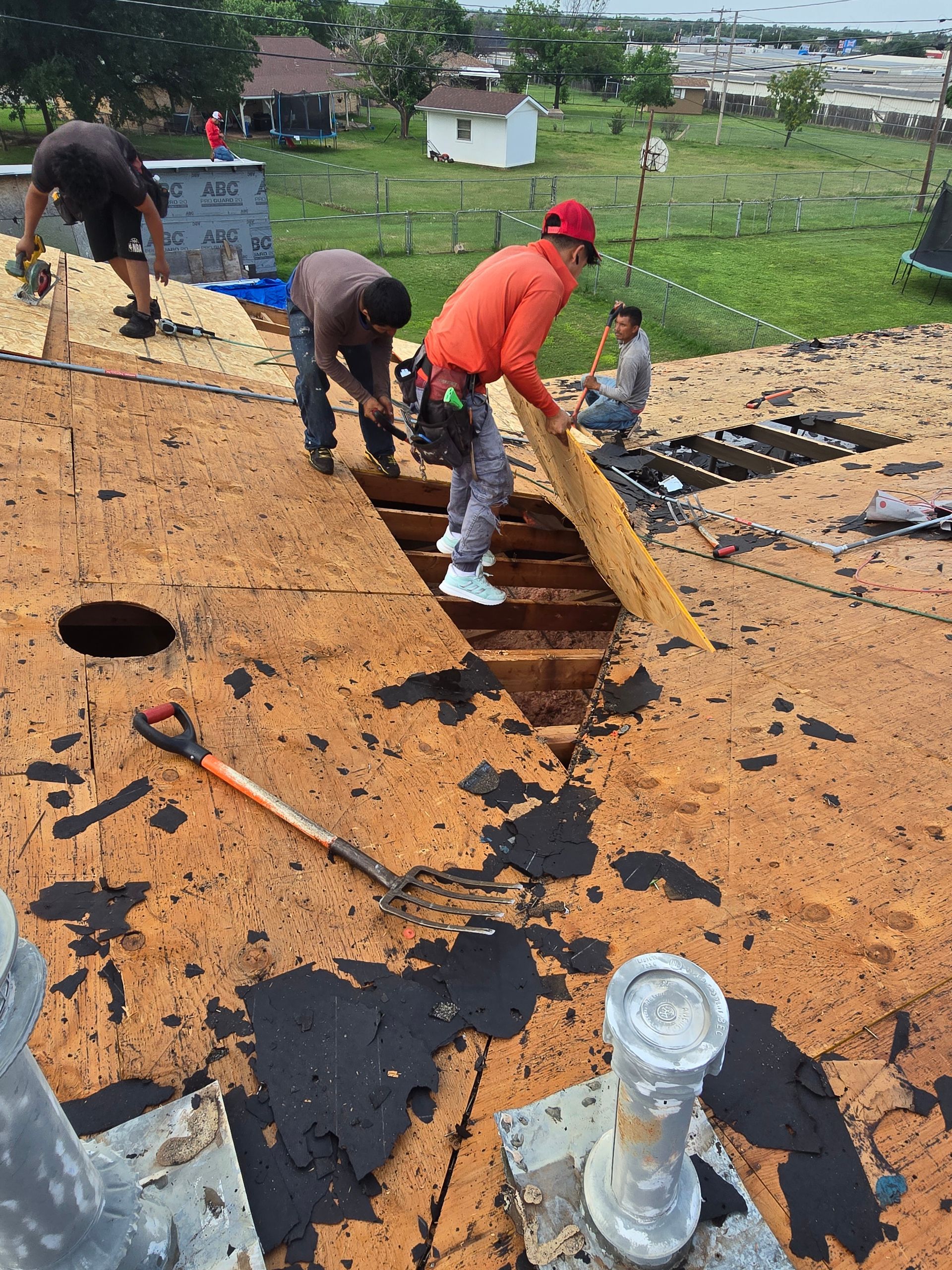 Roofers repairing a shingle roof with black tar paper exposed, using tools near an open section.