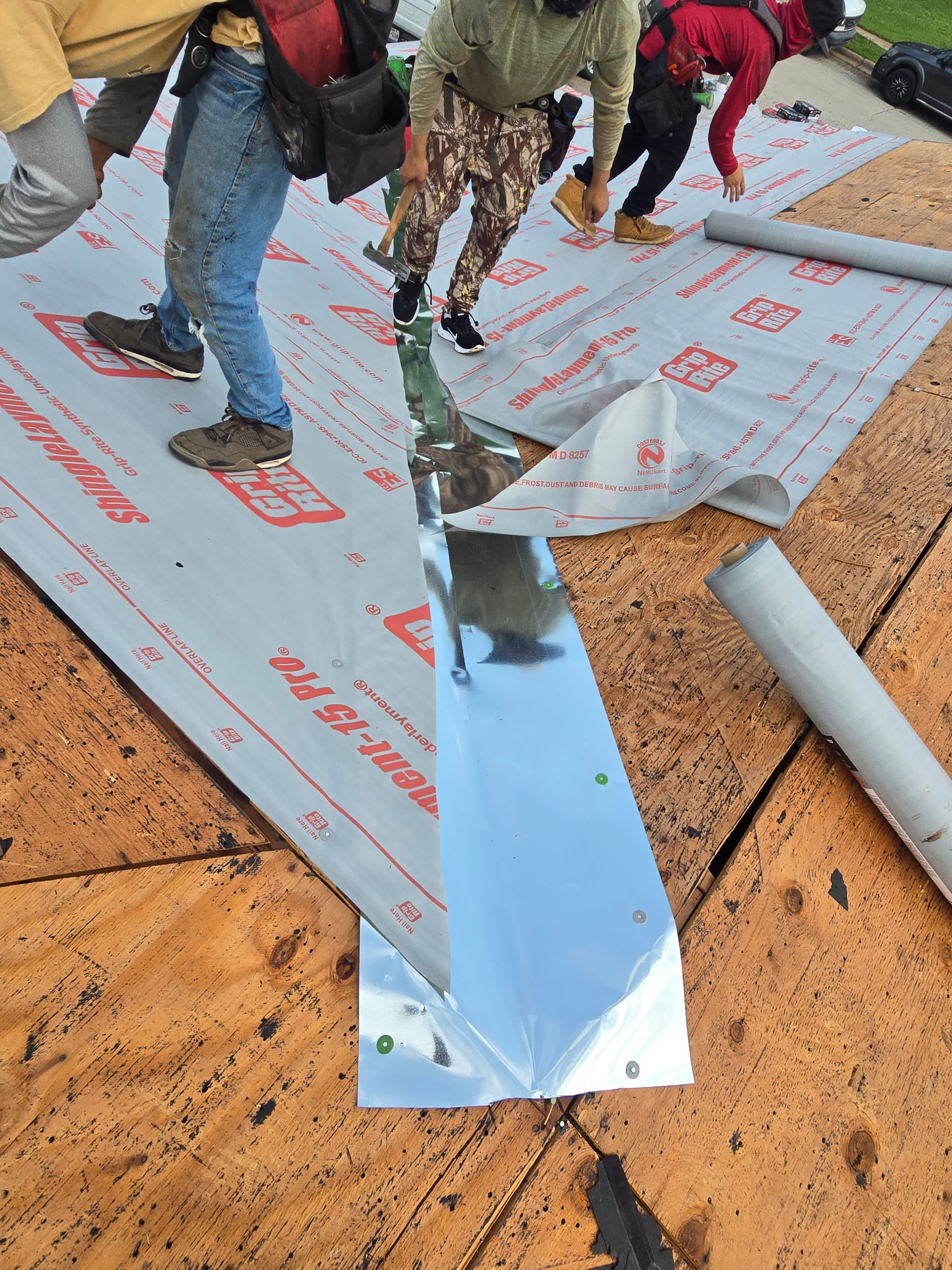 Workers installing metal flashing on a sloped roof with underlayment and exposed plywood.