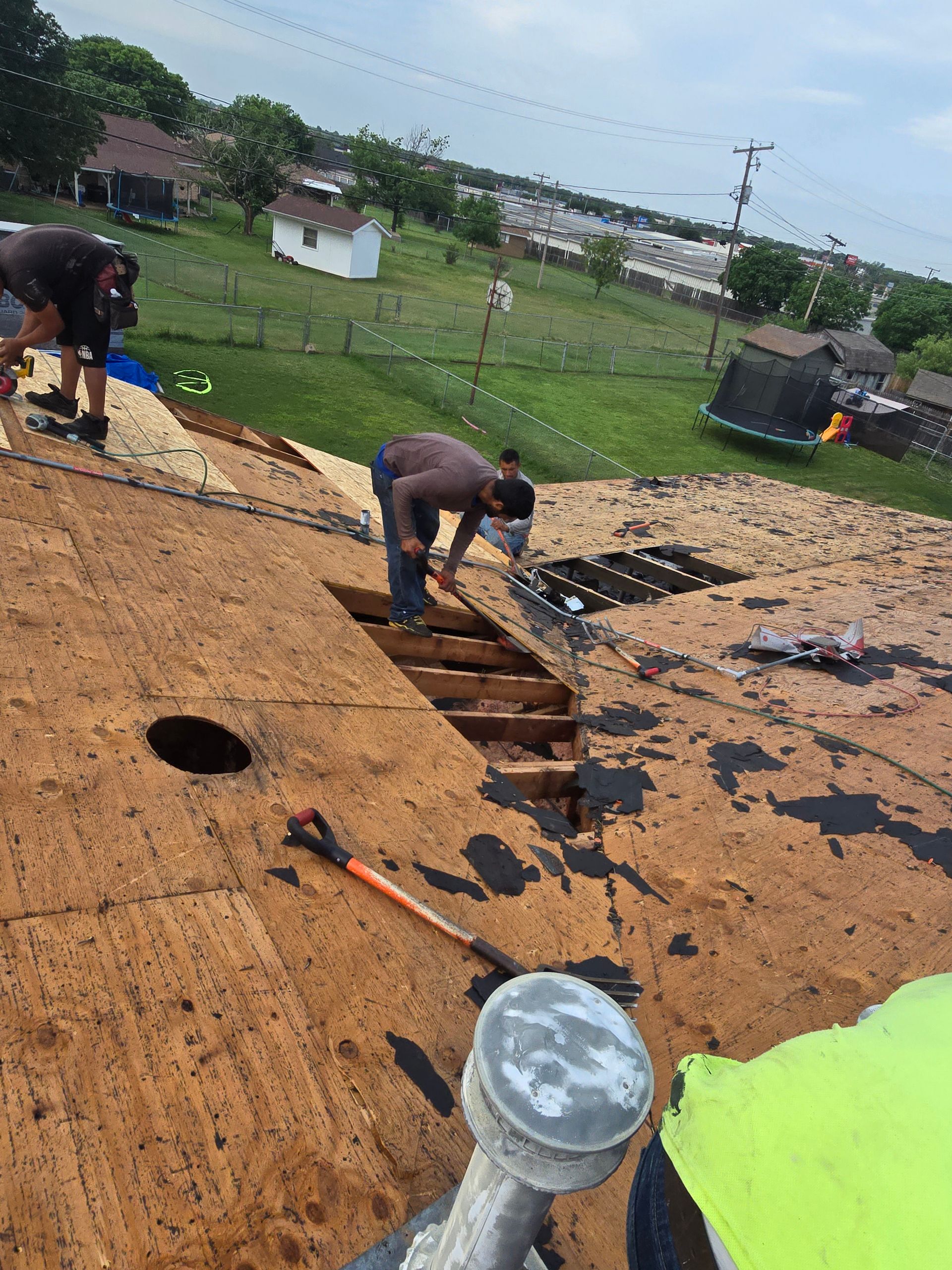 Workers repairing a roof, removing shingles around an open section in a grassy rural area.