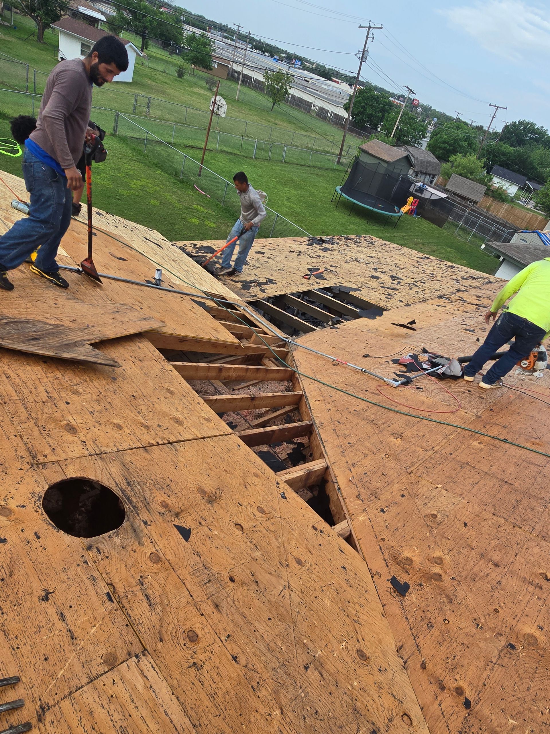 Workers repairing a damaged roof with exposed wooden beams and missing panels in a grassy area