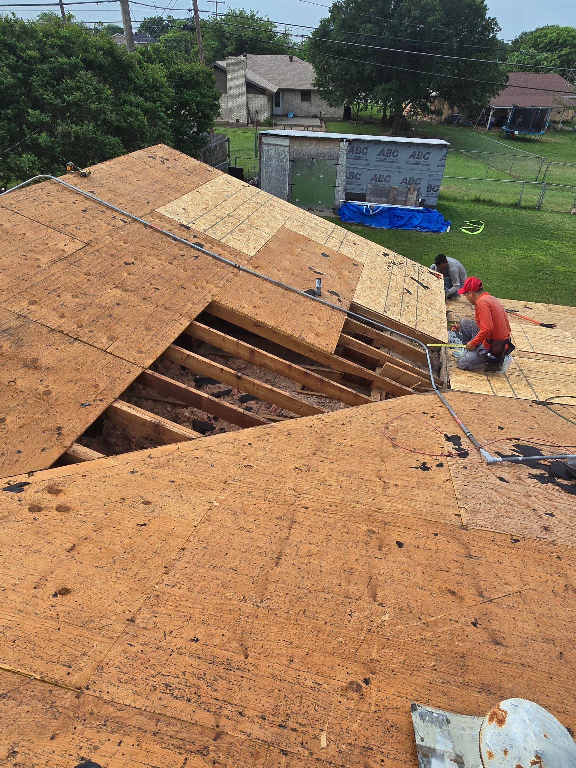 Collapsed wooden roof with exposed rafters in a yard; a person crouches nearby beside a blue barrel.
