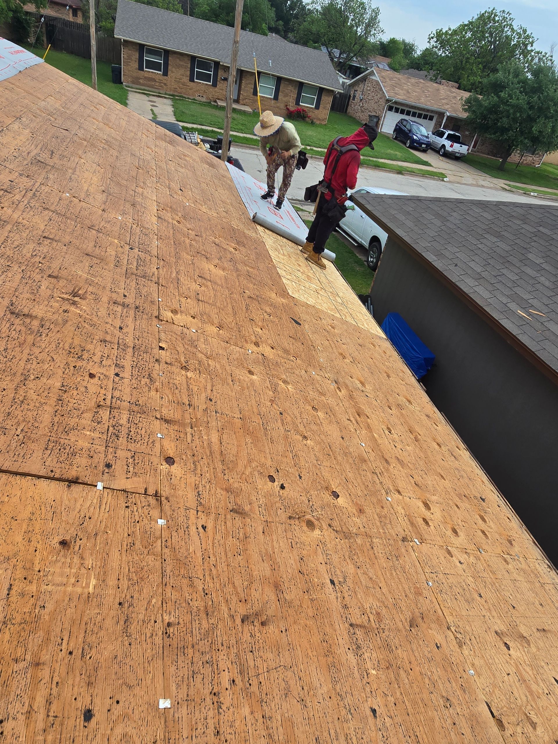 Workers on a steep roof replacing shingles with plywood, houses visible in the background.