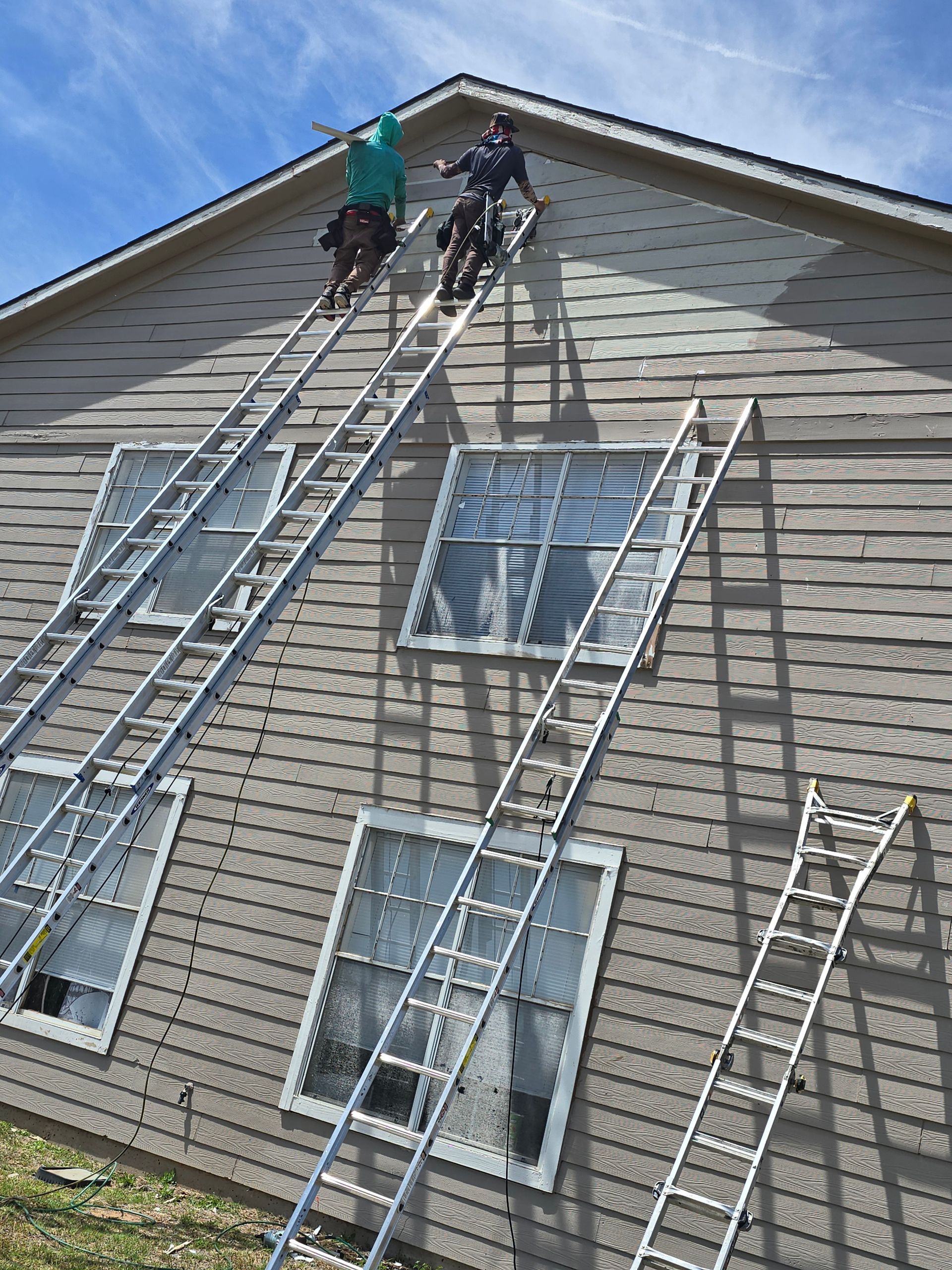 Workers on ladders cleaning the side of a beige house near windows and roofline