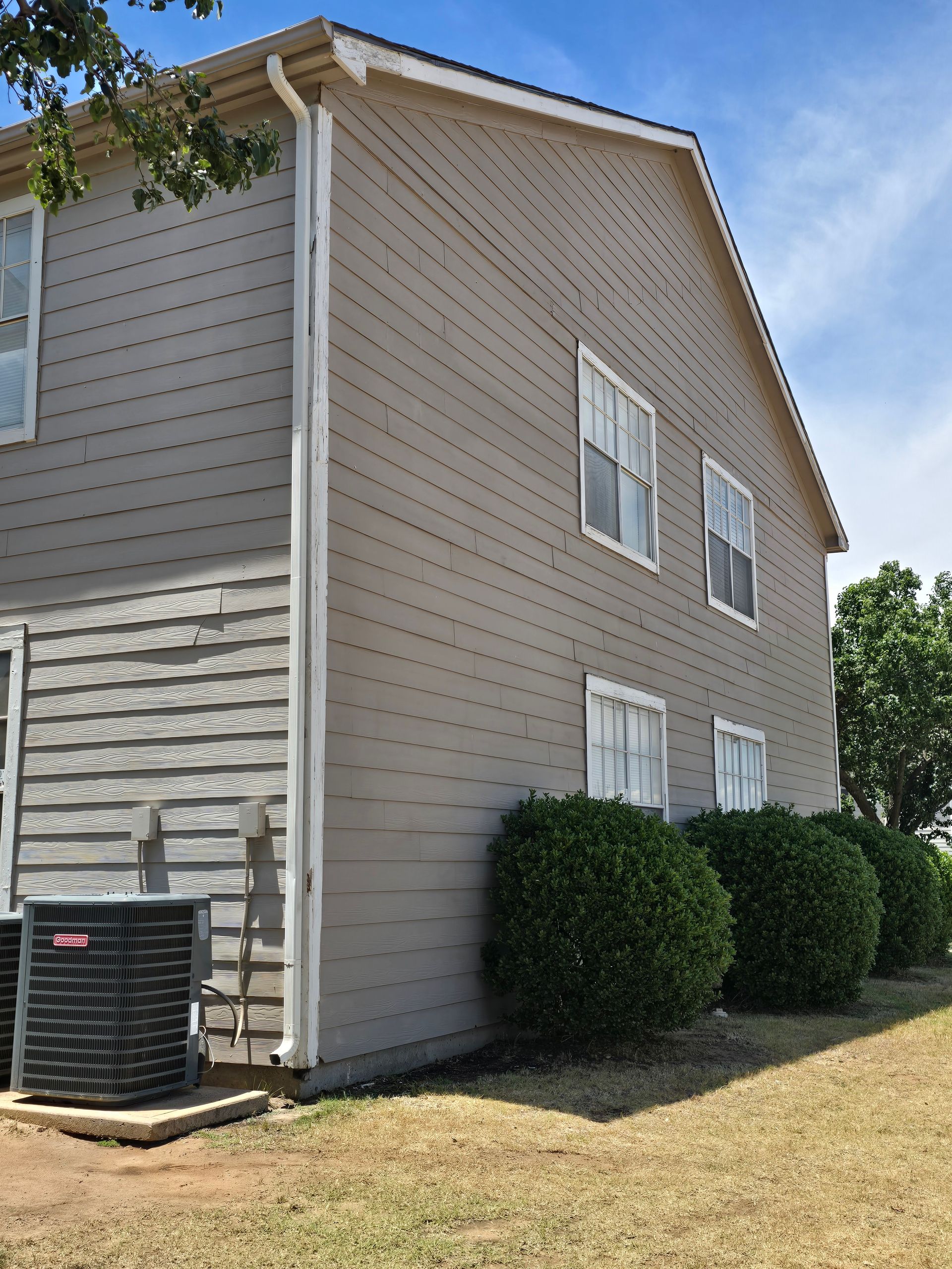 Side view of a beige two-story house with white trim, trimmed bushes, and an air conditioner unit.