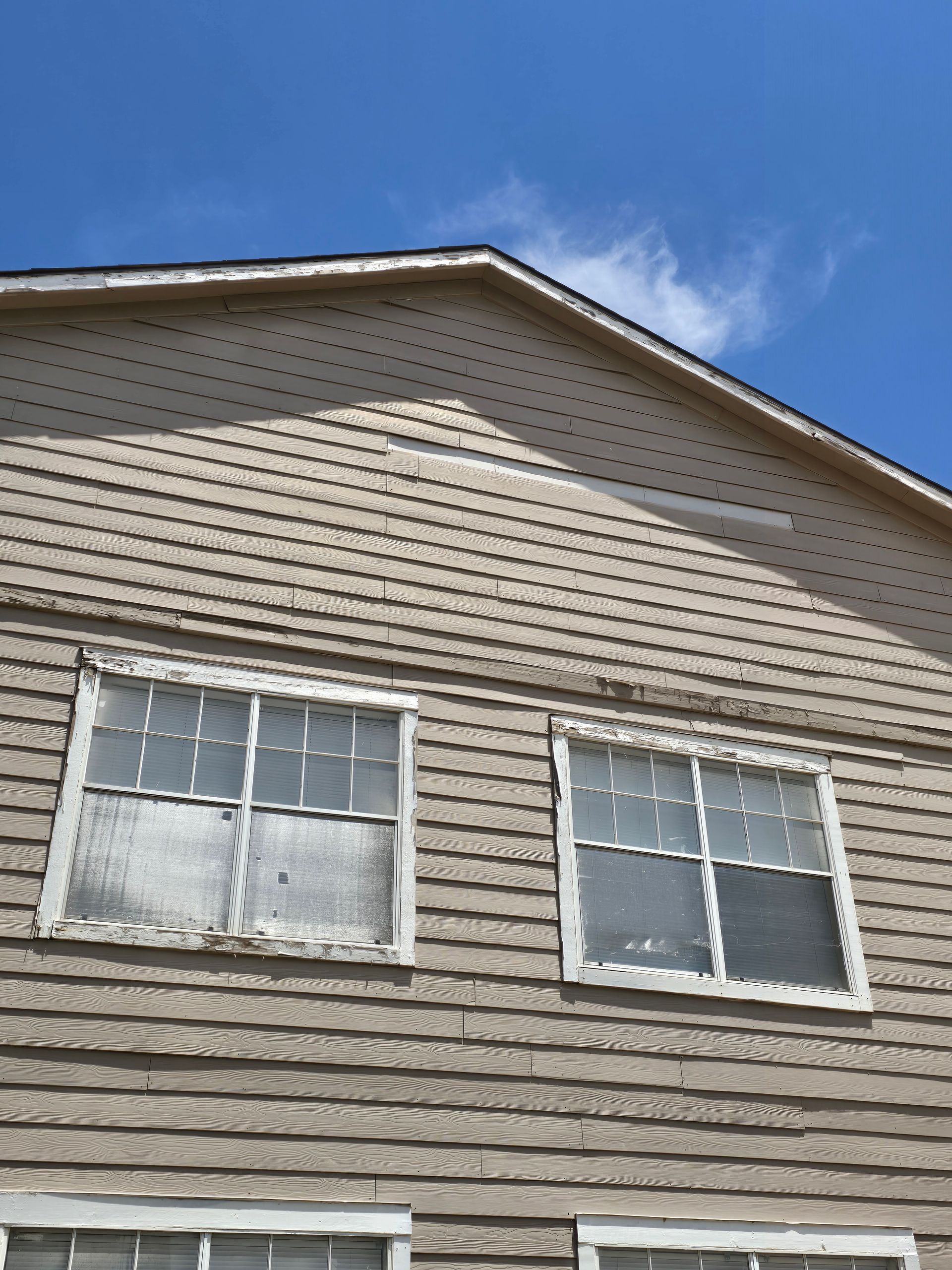 Exterior of a tan house with two white-framed windows beneath a gable roof against a blue sky