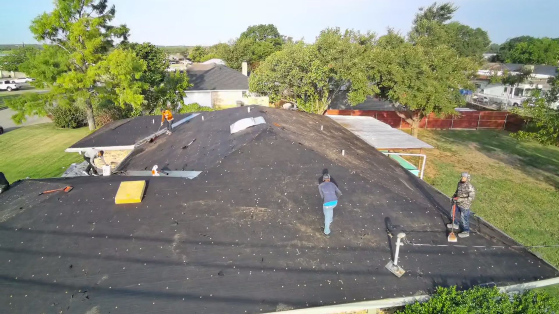 Two workers on a roof under repair, with roofing materials and trees in the background.