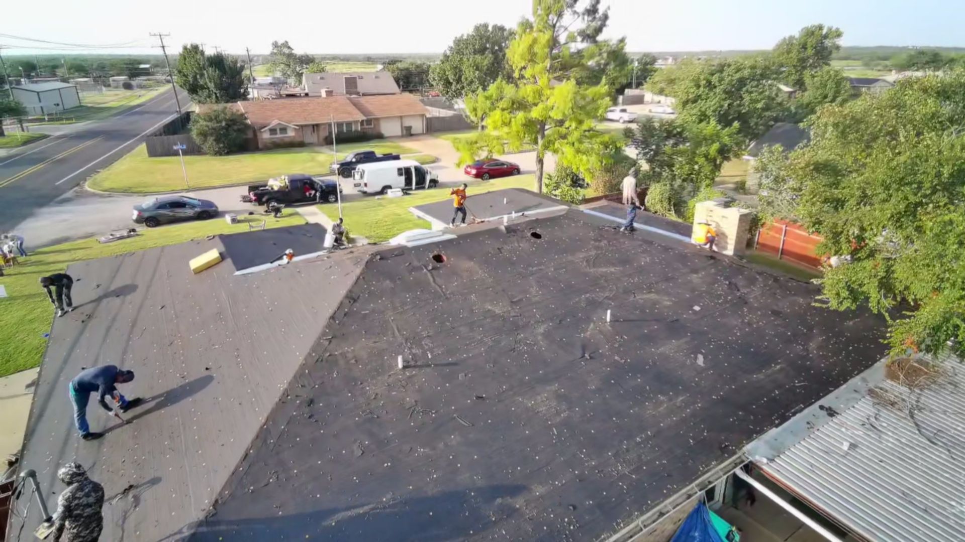 Workers on a rooftop replacing a dark shingle roof in a suburban neighborhood