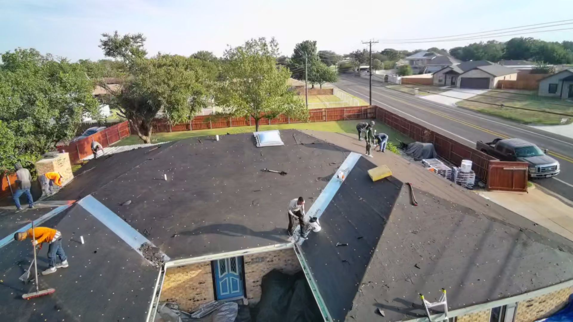 Rooftop under construction with workers, ladders, and materials beside a suburban street.
