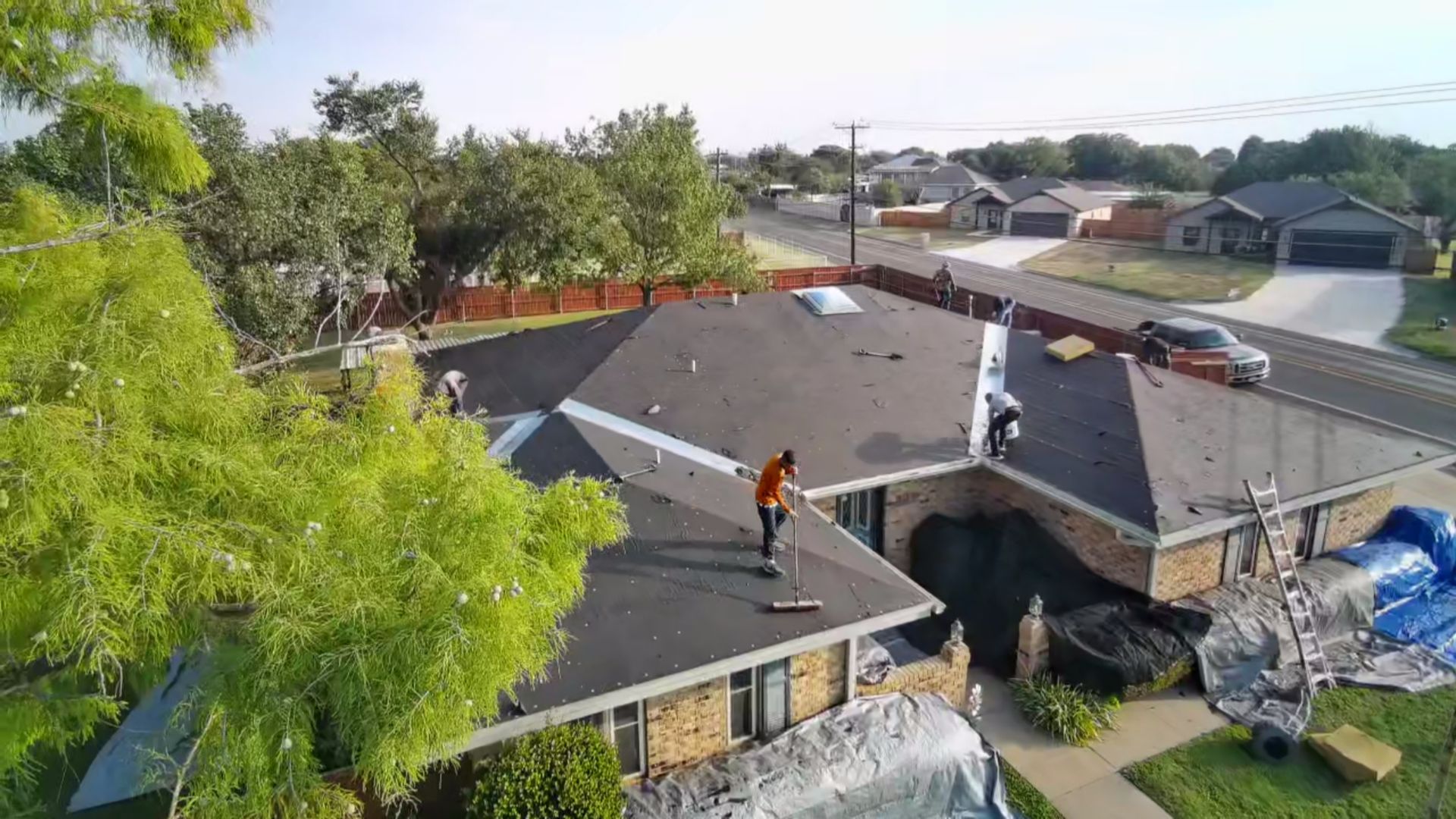 Roofer working on a residential roof in a suburban neighborhood, with trees and nearby houses in view.