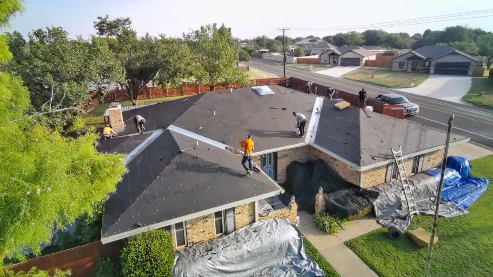 Roofers repairing a house roof with ladders, blue tarp, and tools in a suburban neighborhood