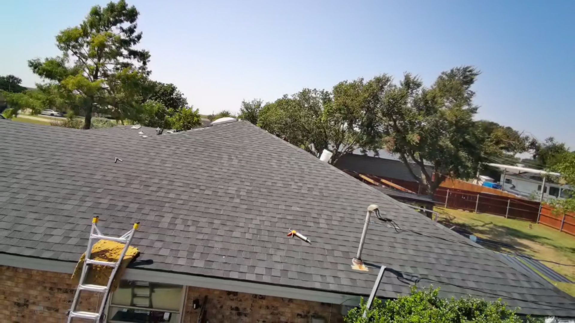 Gray shingled roof under blue sky with trees and a fenced yard in the background