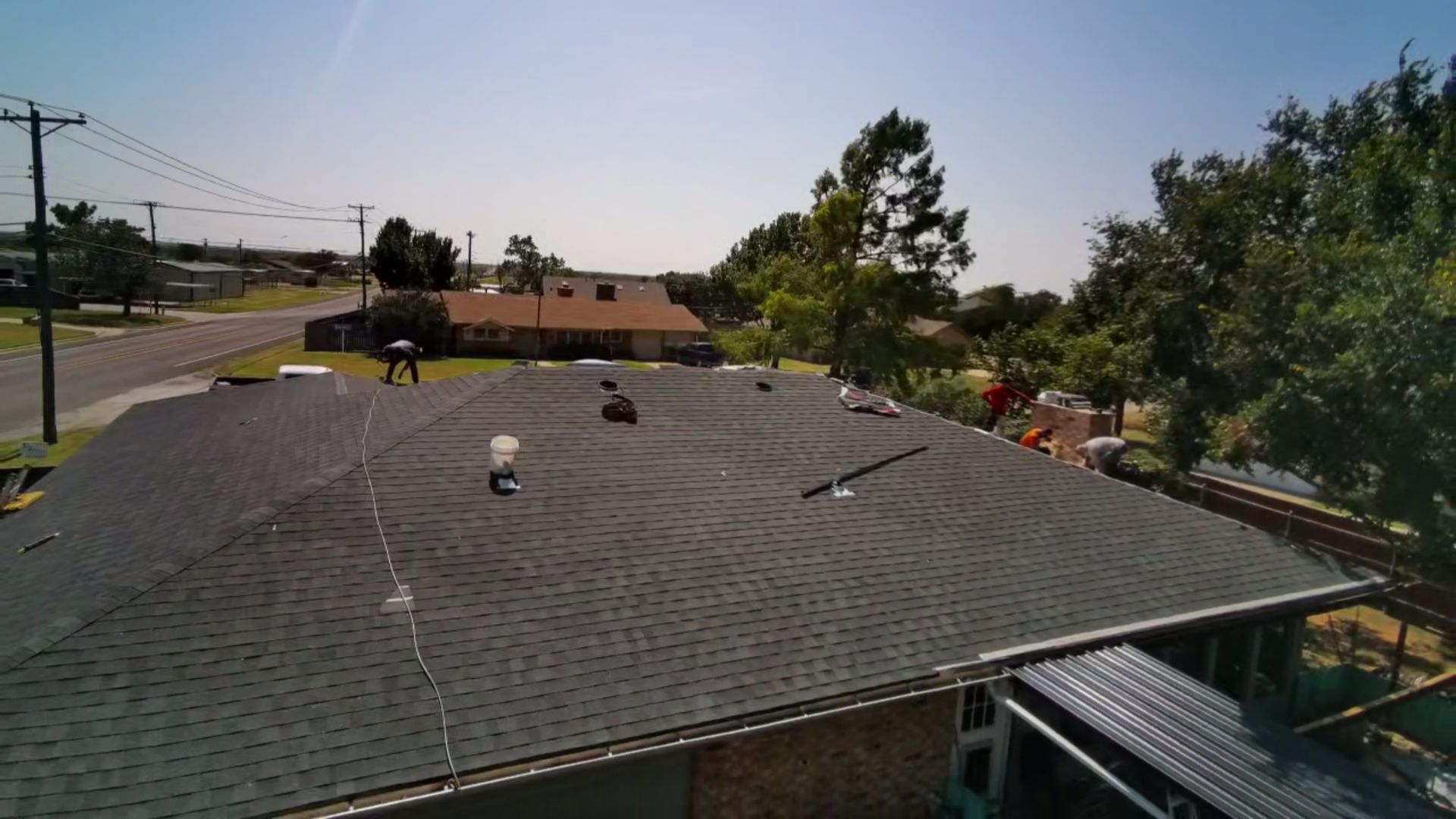 Rooftop with gray shingles and vents, seen from above beside trees and a street under a clear sky