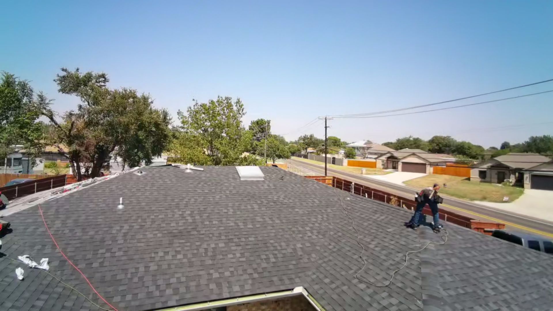 Rooftop view with dark shingles, two workers, and houses and trees in the background.