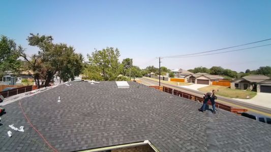 Rooftop view with dark shingles, two workers, and houses and trees in the background.