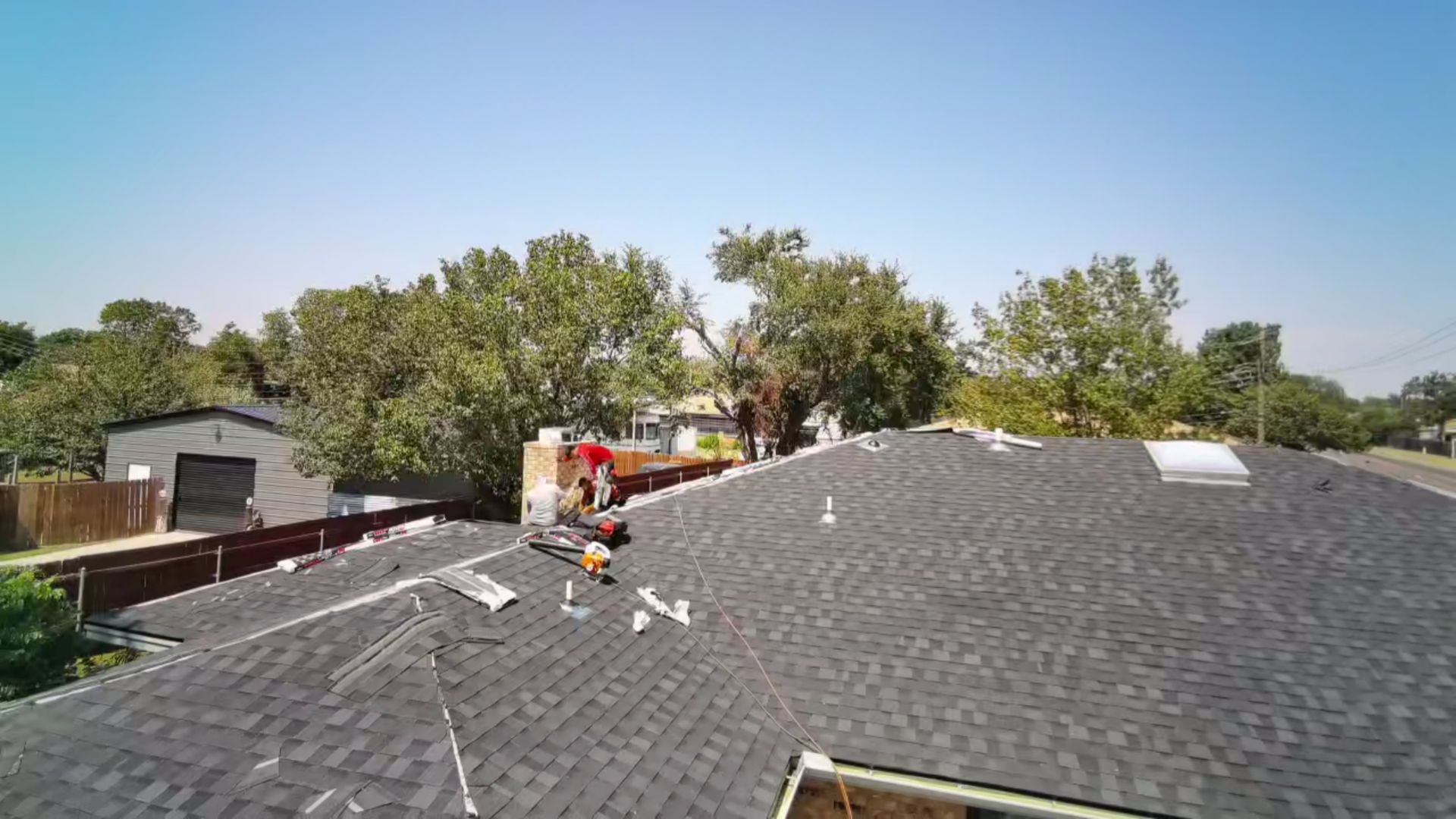 Roofers working on a shingled roof with trees and a house in the background