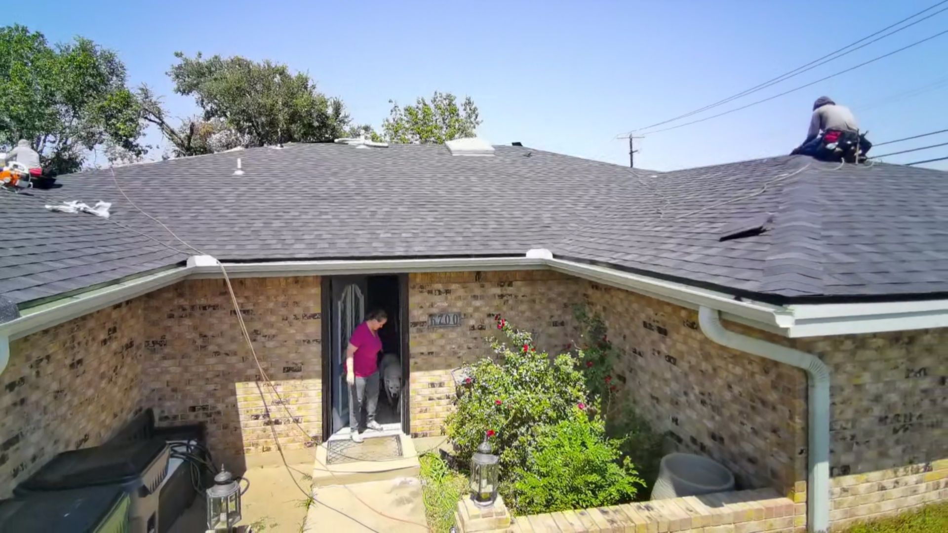 Brick house roof with worker kneeling near the ridge and a person standing in the doorway below
