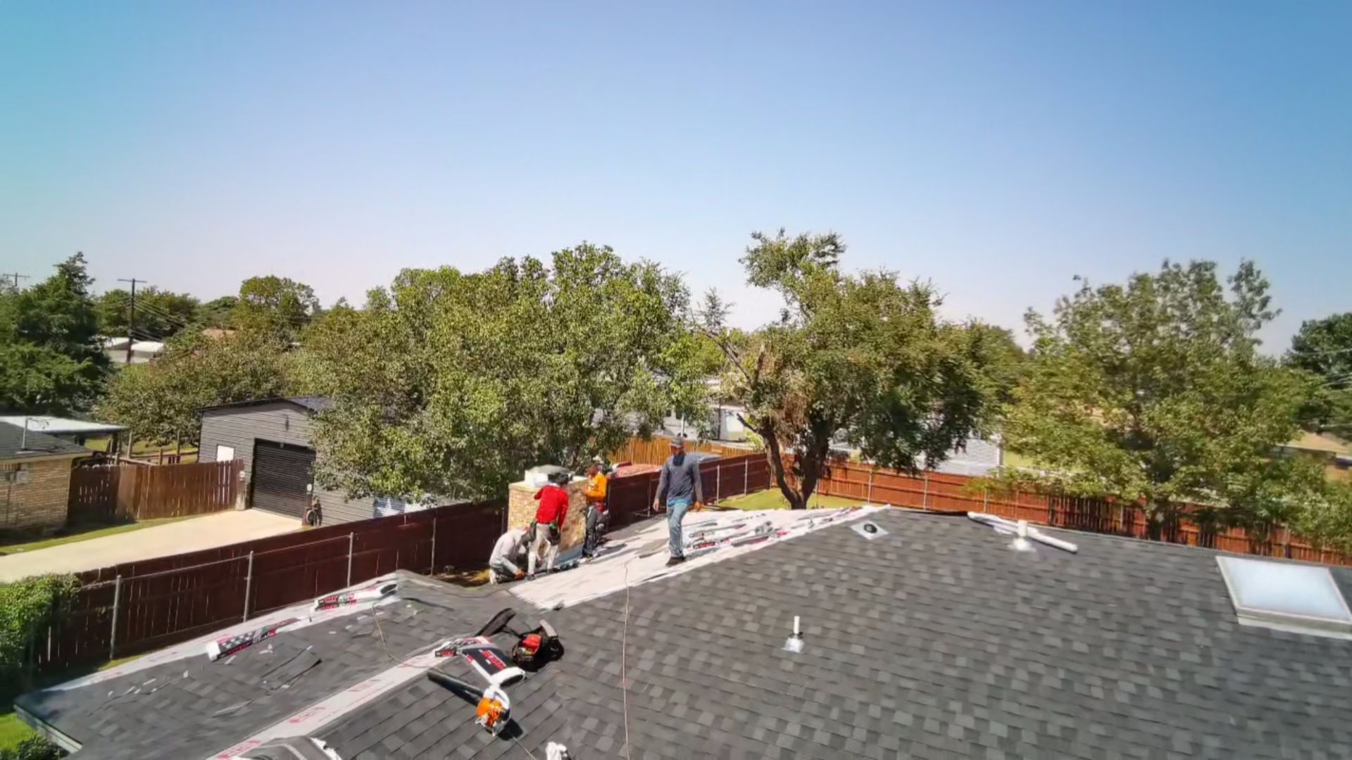 Workers repairing a dark gray shingle roof beside trees and houses on a sunny day