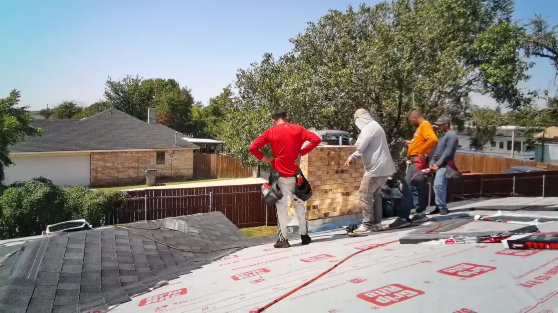 Workers installing roofing on a house, with red and orange shirts on a sunny day.