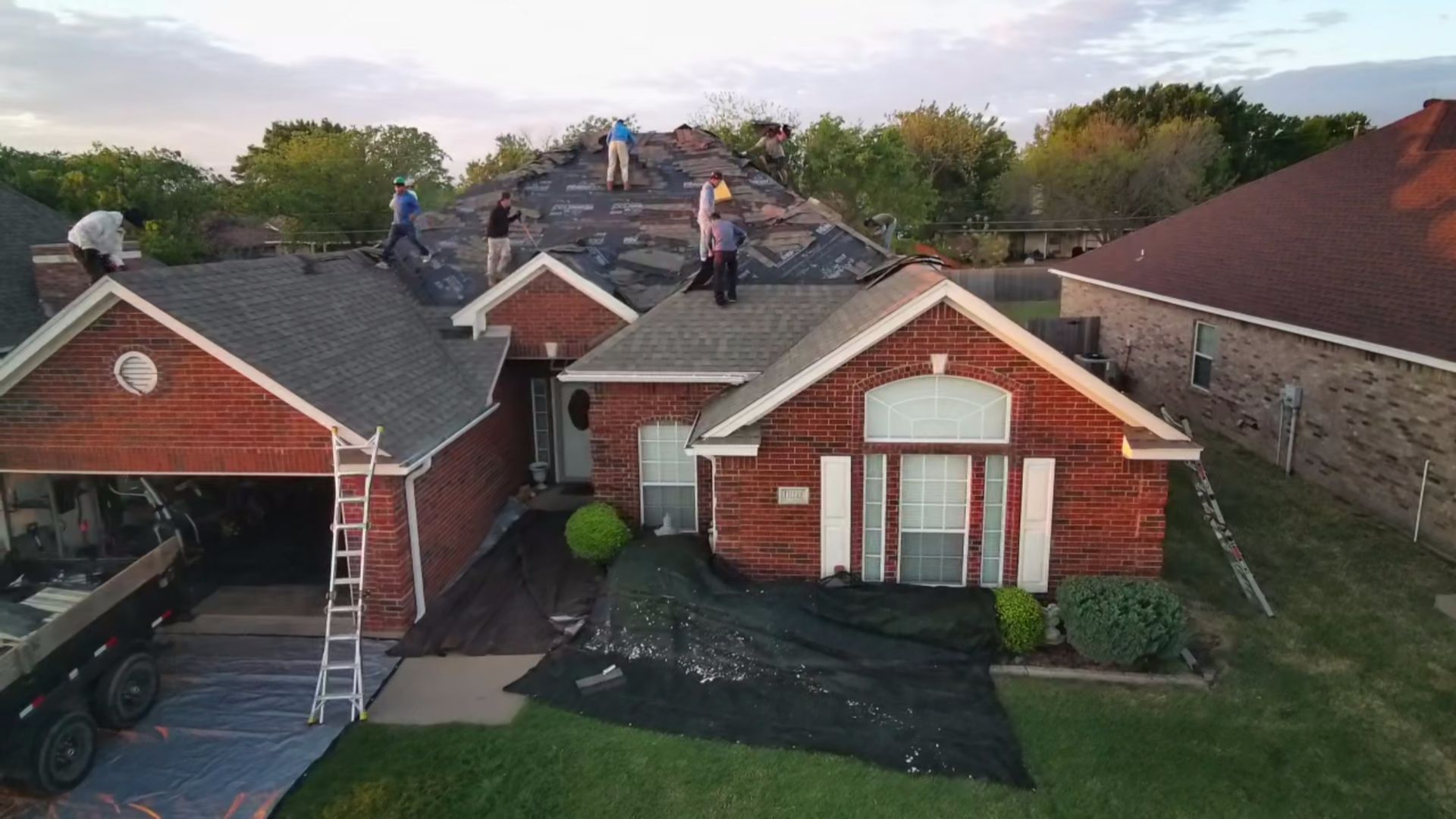 Two brick houses with dark roofs, ladders, and roofing materials, viewed from above in a suburban yard.