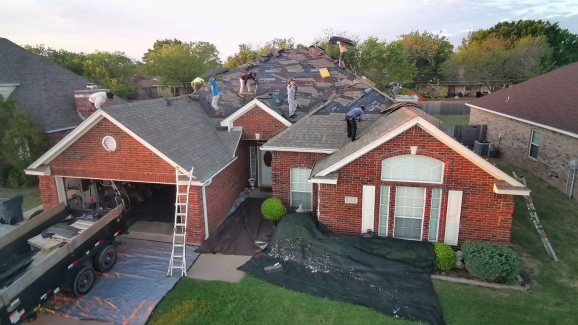 Aerial view of two red brick houses with roof work and a ladder in a suburban neighborhood.