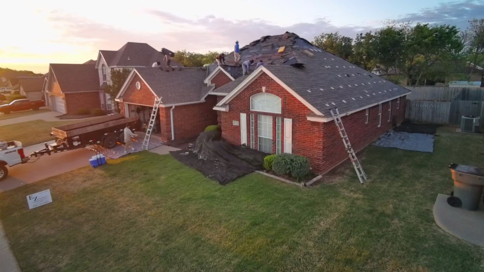 Aerial view of brick suburban houses and lawns at sunset, with a driveway and fenced backyard.