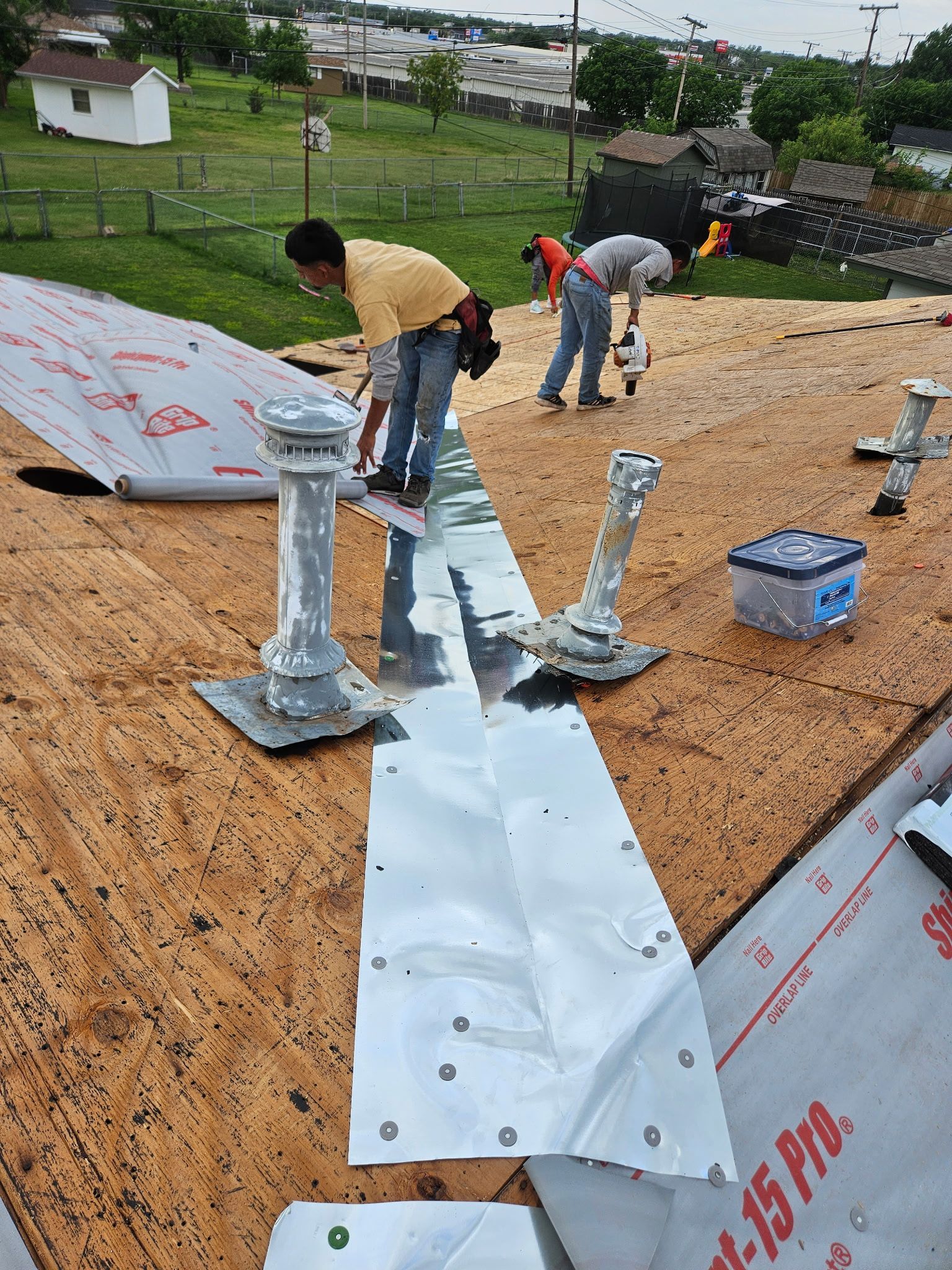 Workers installing roof flashing on a sloped roof with silver underlayment and gray vents.