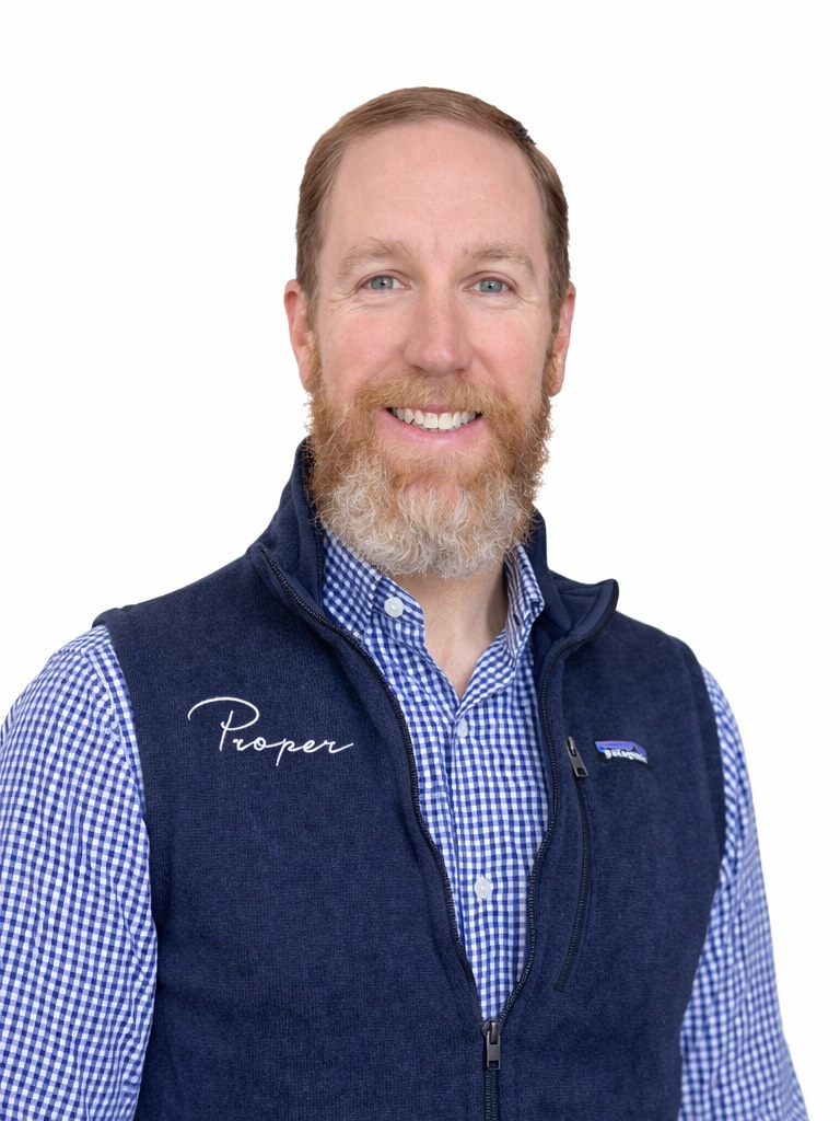 Smiling man with a red beard wearing a blue plaid shirt and navy vest with