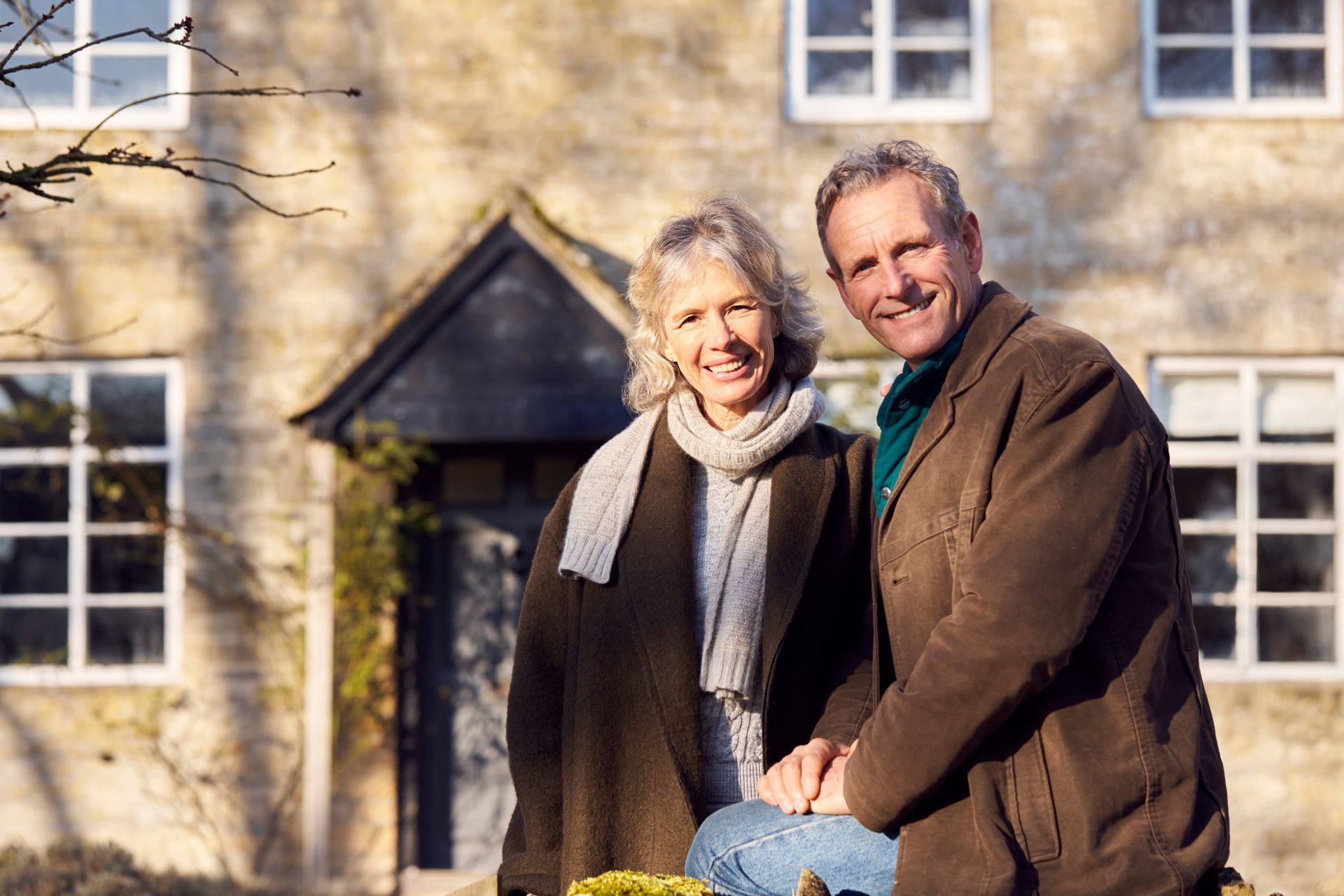 Smiling couple standing outside a house in winter light, with a stone wall and windows behind them