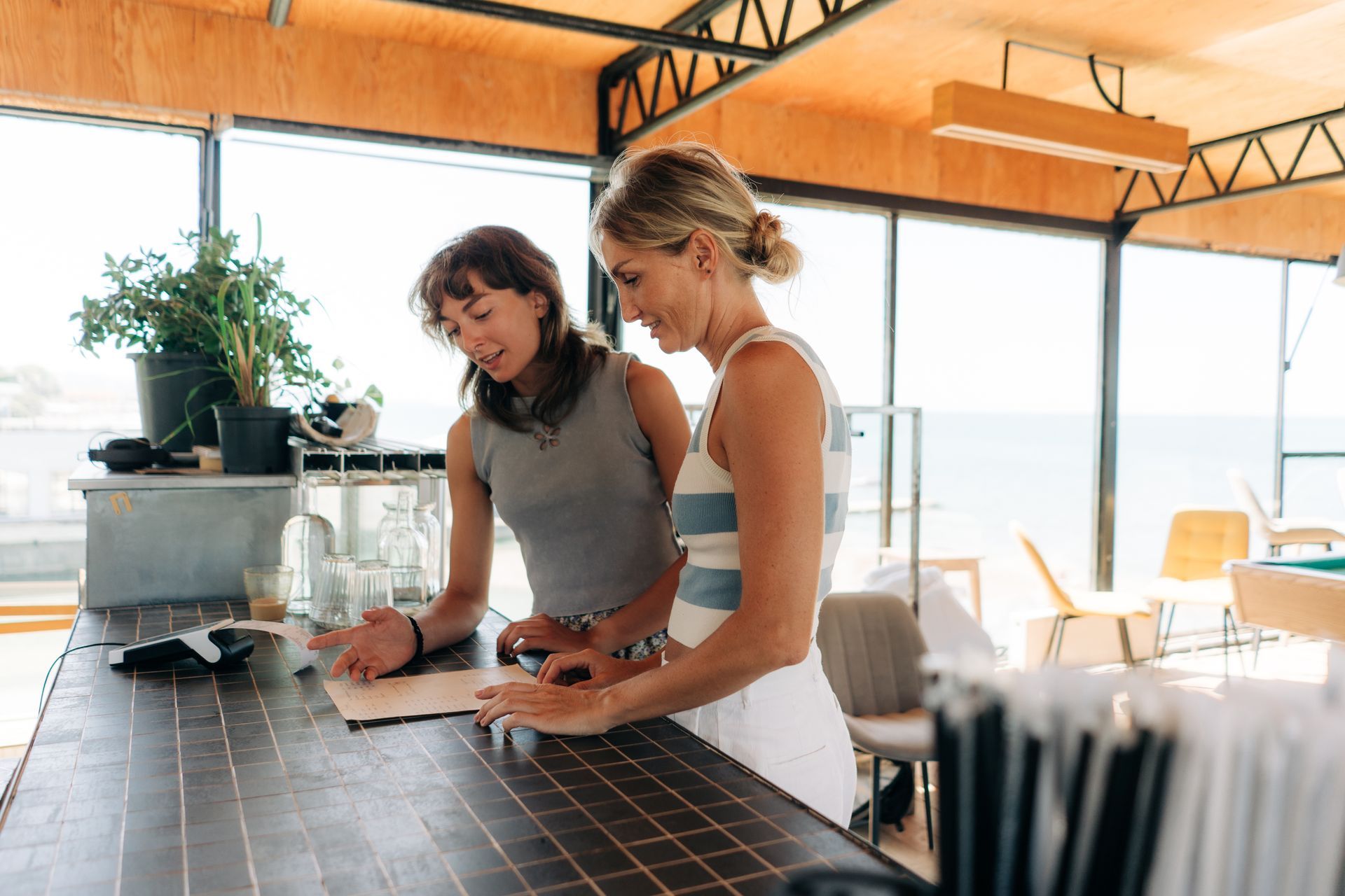 Two women preparing food on a tiled counter in a bright kitchen with large windows.