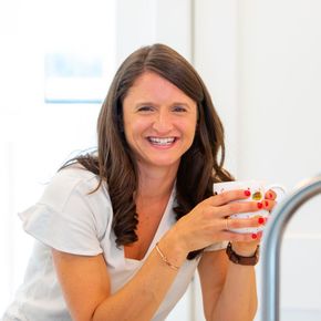 Woman in white top smiles, holding a mug, standing near a sink.