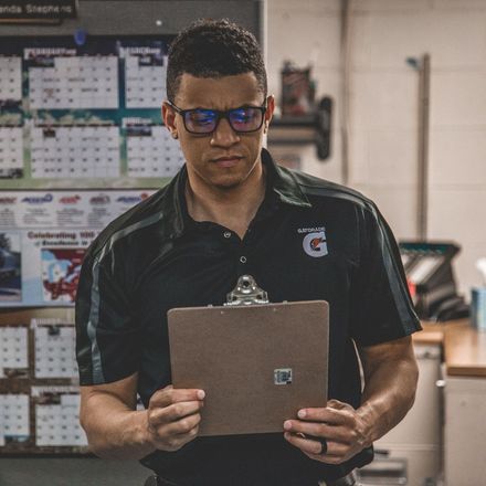 Man in glasses, holding a clipboard, looking down, standing in front of a calendar.