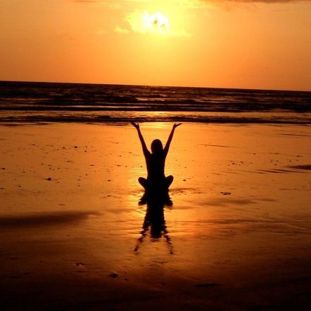 Person sitting on beach, arms raised, silhouetted against the sunset.