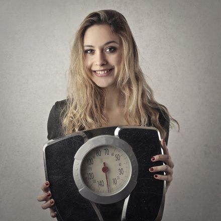 Woman smiling, holding a black bathroom scale, weight goal, looking at viewer, light background.