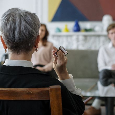 Counselor, seated with back to camera, holds glasses, facing couple in therapy session.