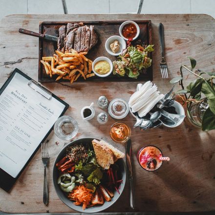 Wooden table with steak, fries, salad, bowl with food, drinks, menu, cutlery.